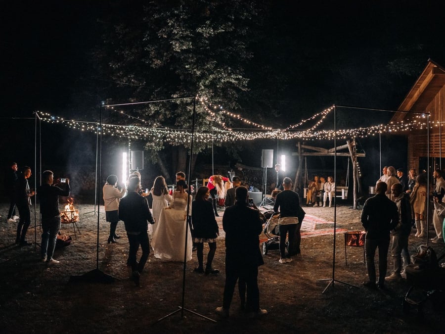 Réception de mariage en extérieur à la tombée de la nuit : guirlandes lumineuses tendues au-dessus des invités, deux braseros allumés, la mariée en robe blanche au centre.