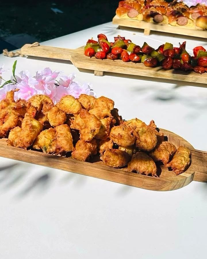 Buffet dressé sur nappe blanche : planches de bois garnies d’accras et de brochettes, fleurs posées entre les planches.