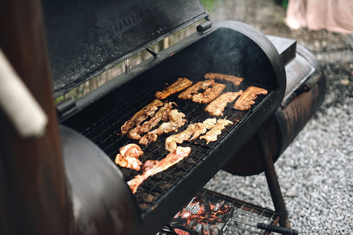 Un smoker à foyer décalé, capot ouvert : des pièces de viande alignées sur la grille et la fumée qui monte.
